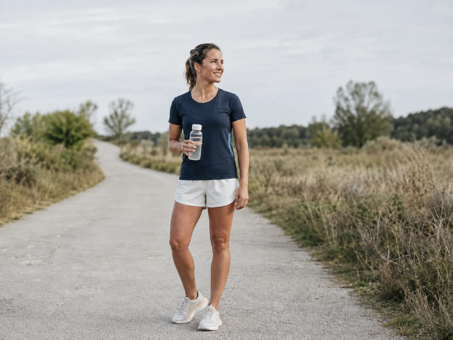Fröhlich lächelnde Läuferin in Navy und Off-White mit einer vorbereiteten Trinkflasche bei einer Pause auf ihrer Strecke; generierte Anwendungsszene